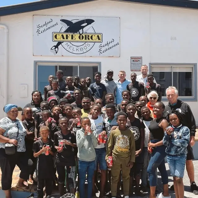 A youth football team and their coaches gathered outside the Café Orca cottage under the orca sign