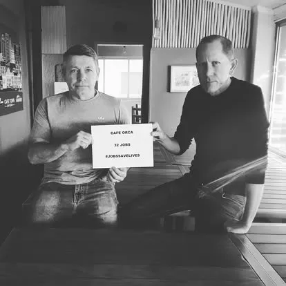 Two men at an empty table holding a sign reading “Café Orca, 32 jobs, #JobsSaveLives”