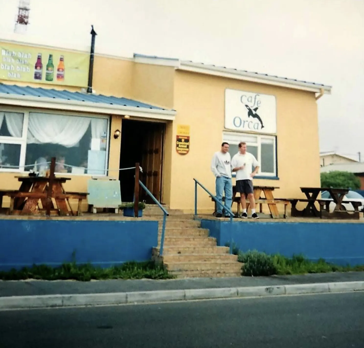 The original Café Orca on Beach Road in 1997: a yellow cottage with a blue wall, picnic tables and two men standing on the stoep