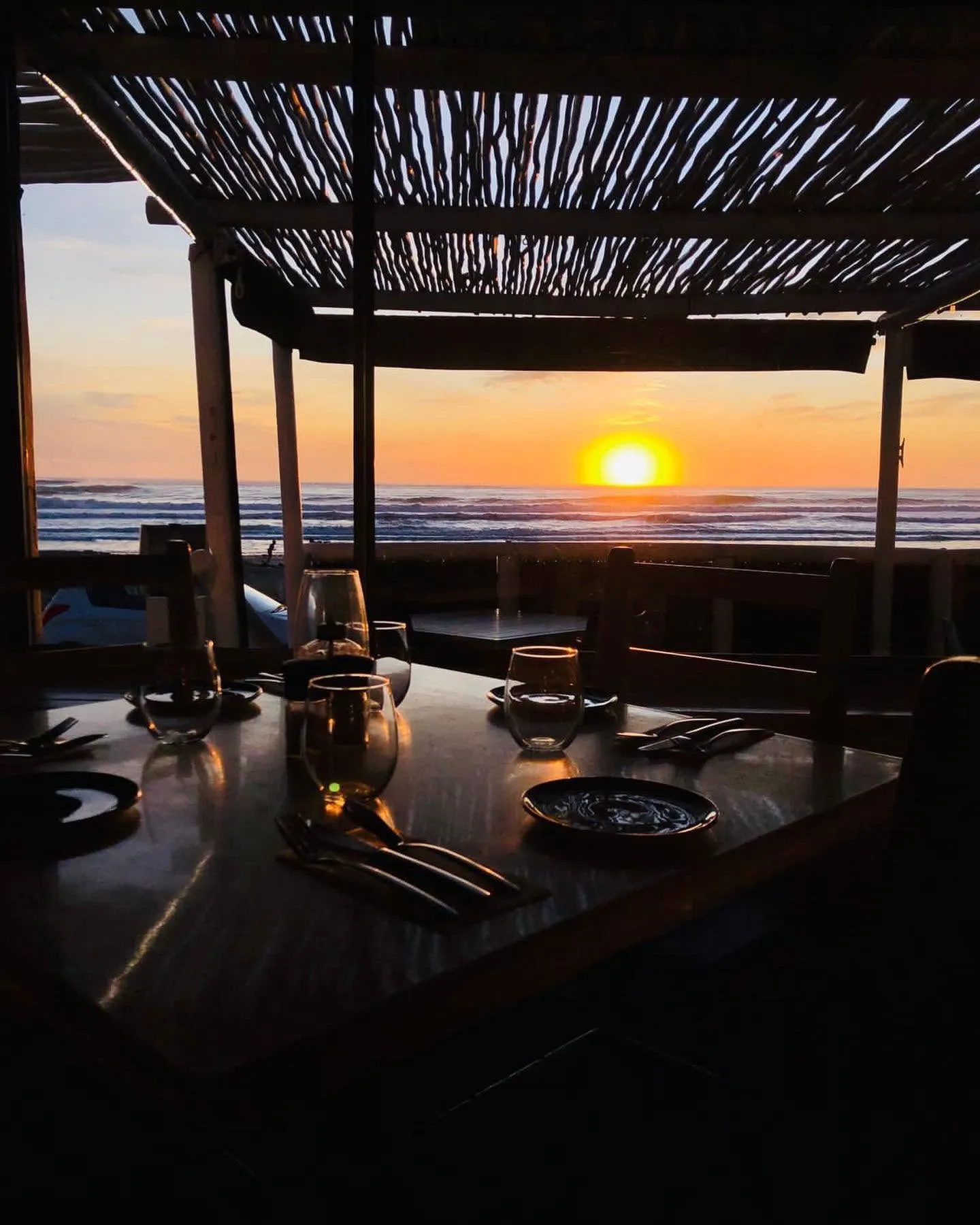 A table set for dinner under the reed roof, the sun setting over the Atlantic beyond